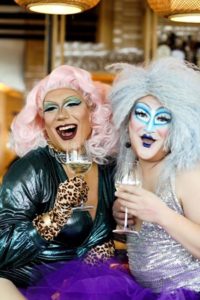 Two drag queens smiling and holding glasses of champagne